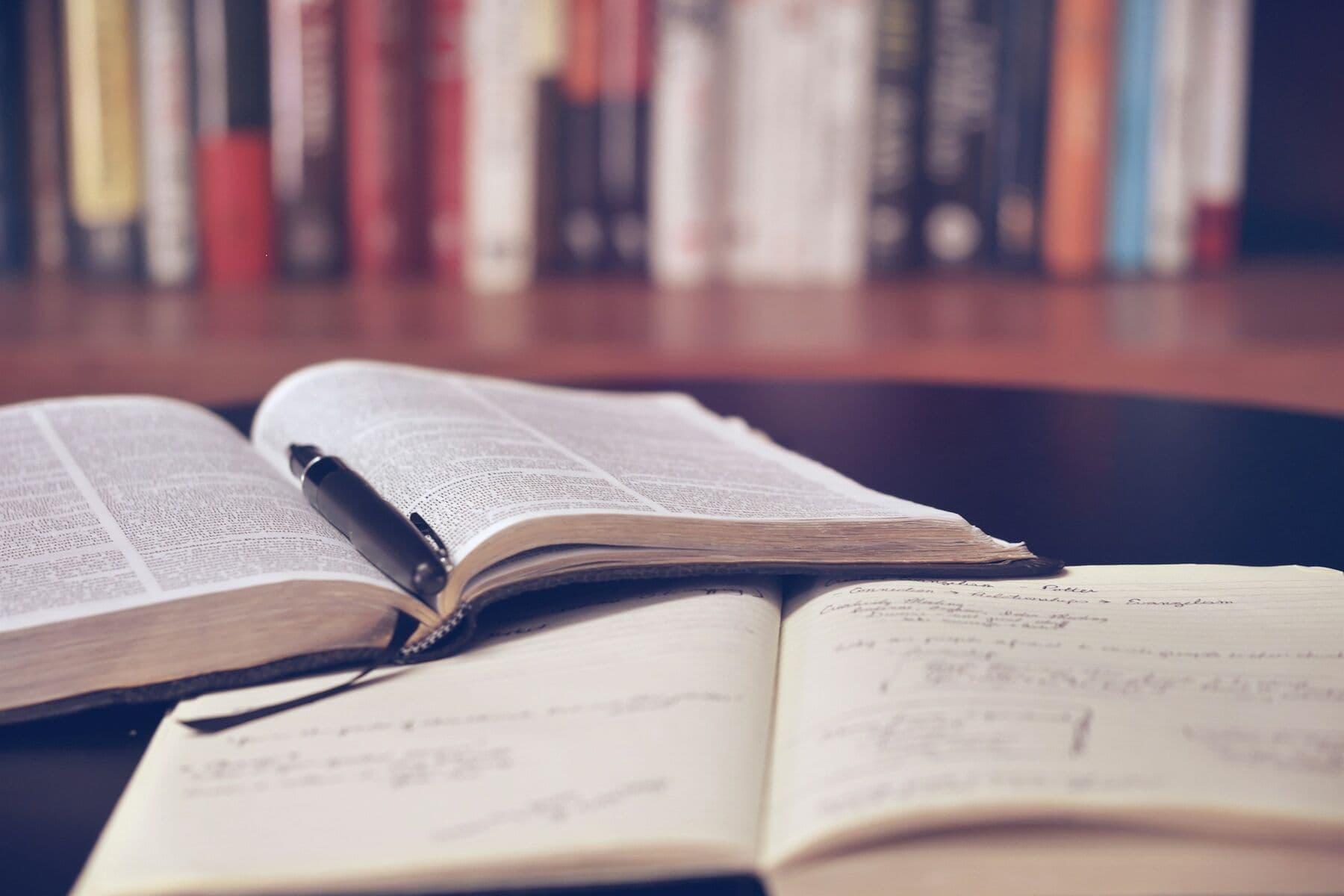 Two open books with a pen on a desk in front of a bookshelf