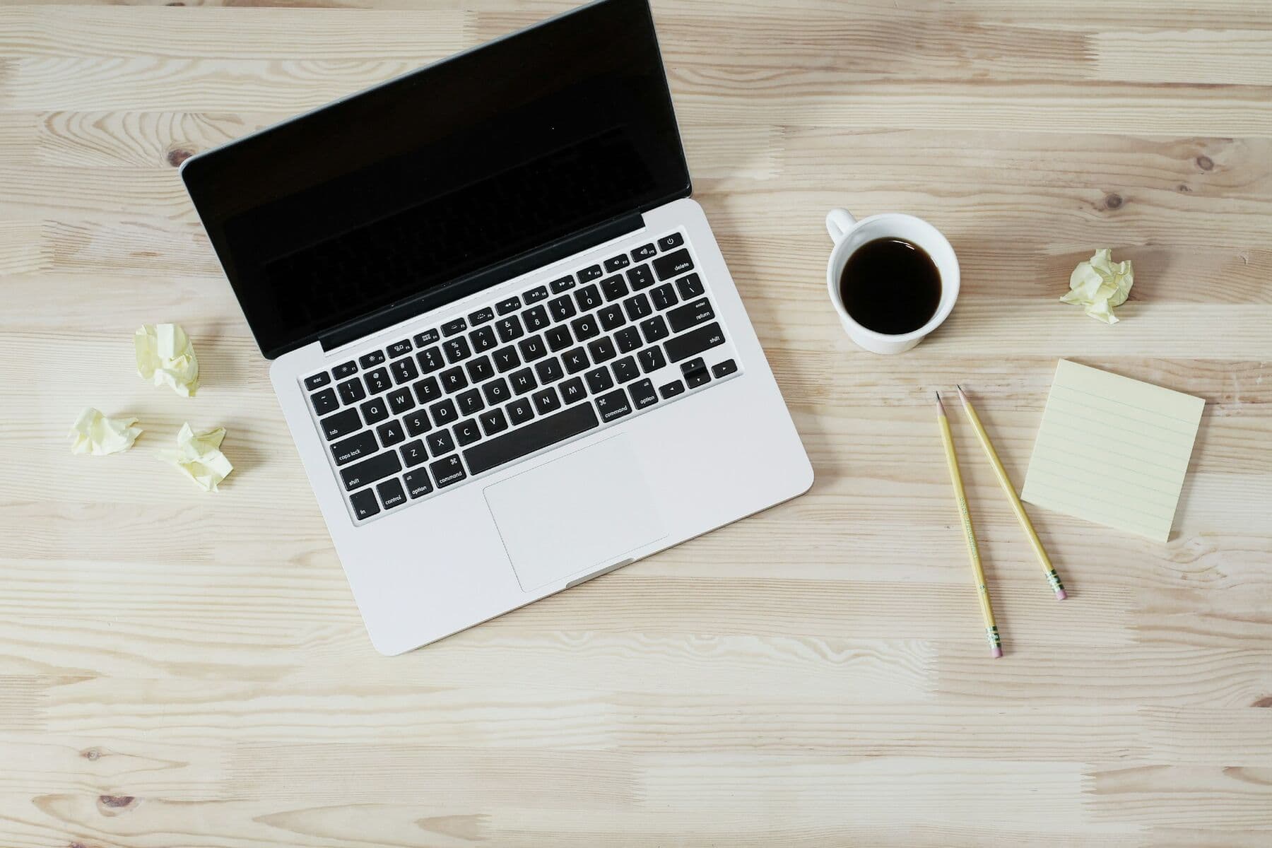 A laptop, coffee, pencils and a notepad on a wooden desk, seen from above